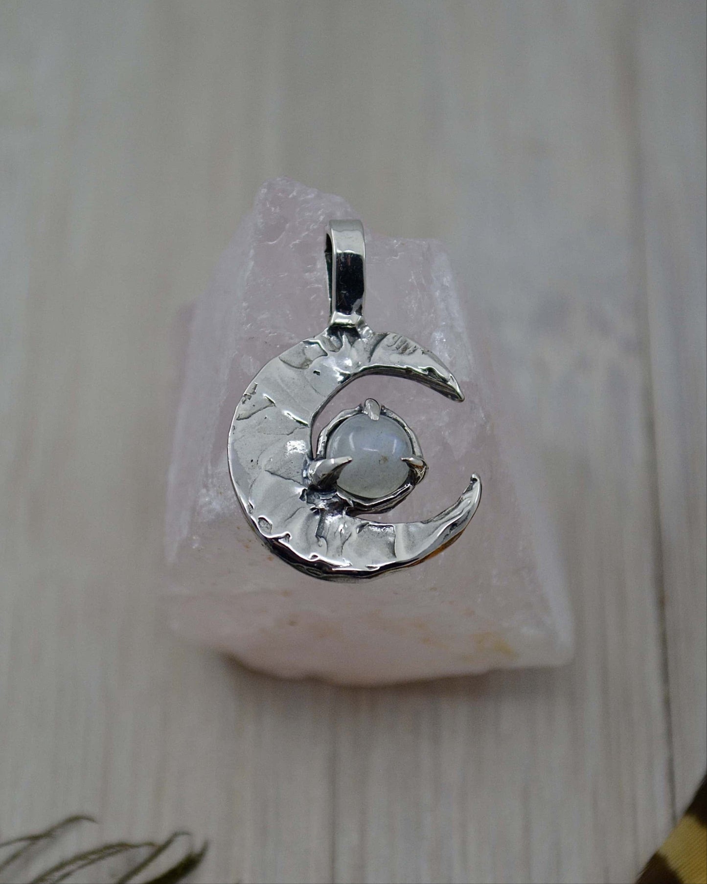 A sterling silver pendant featuring a crescent moon design with a Rainbow Moonstone in the center, displayed on a pink crystal stand.