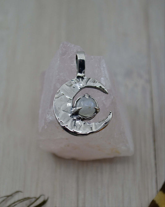 A sterling silver pendant featuring a crescent moon design with a Rainbow Moonstone in the center, displayed on a pink crystal stand.