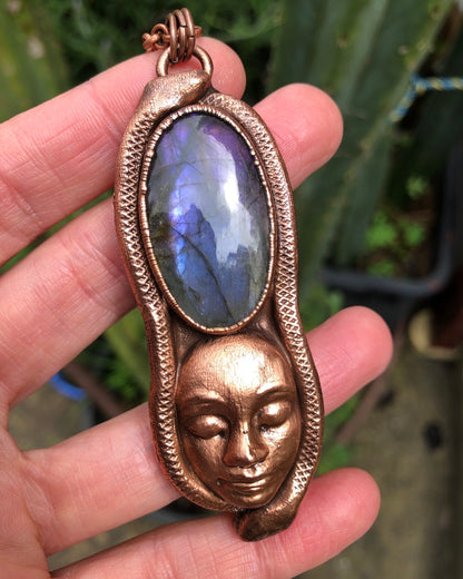 copper pendant with a purple labradorite, a goddess face below it and two snakes along the outer edges, electroformed