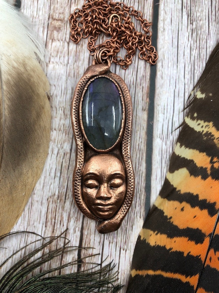 copper pendant with a purple labradorite, a goddess face below it and two snakes along the outer edges, electroformed